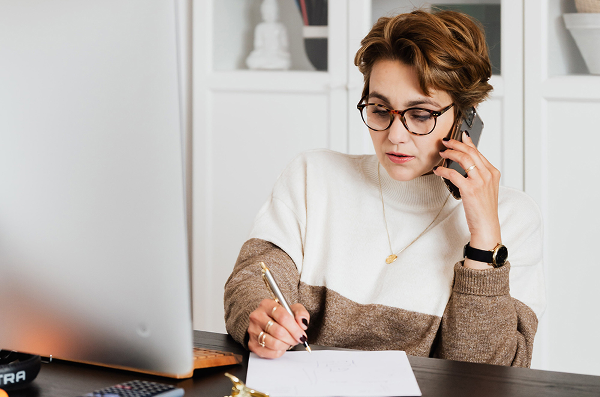 Photographie d'une femme aux cheveux courts et avec des lunettes et un pull bicolore blanc et marron. Celle-ci est au téléphone et écrit en même temps sur une feuille.
