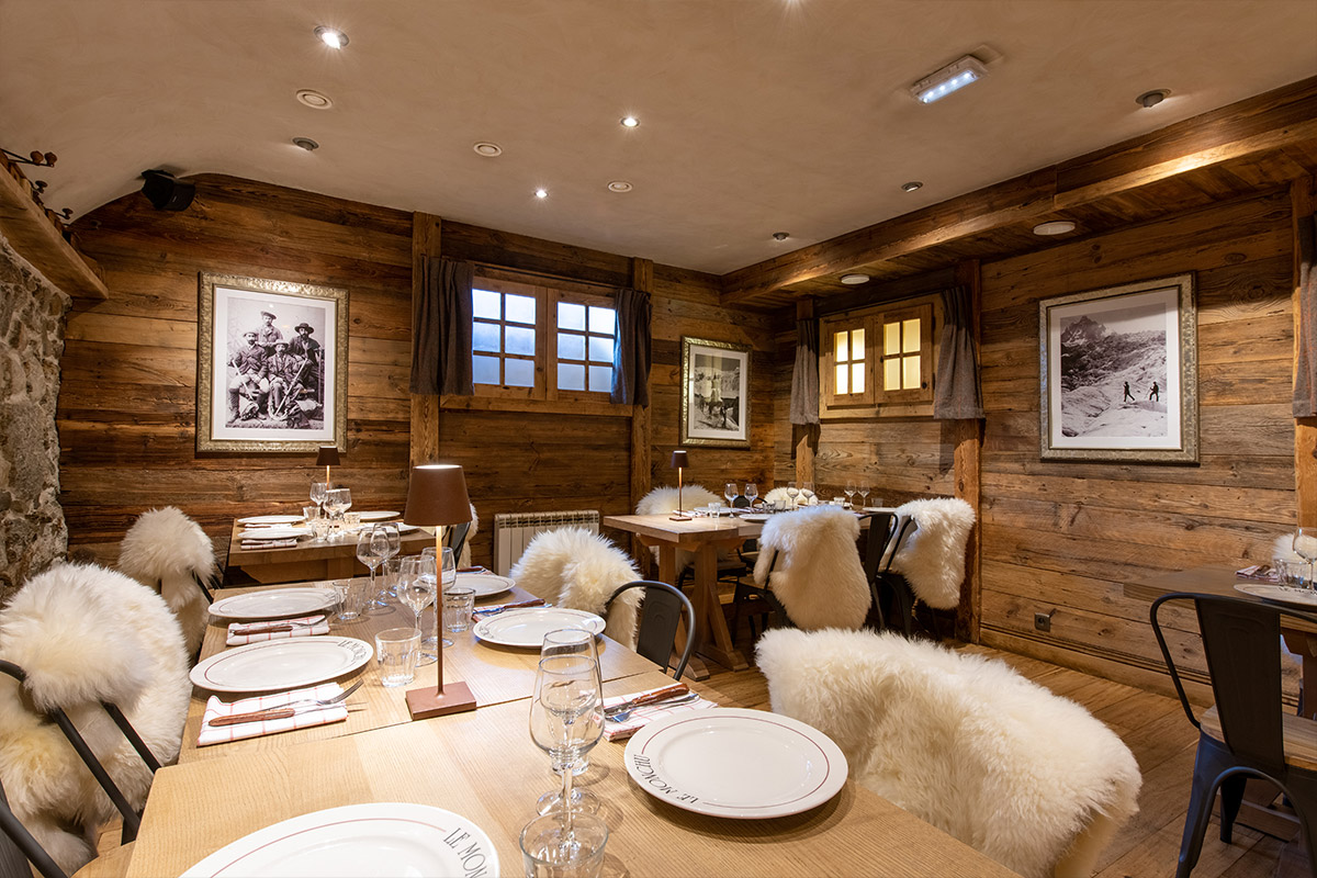Photographie de la salle intérieur du restaurant Le Monchu situé à Chamonix, dans la vallée du Mont-Blanc. Les murs et les tables à manger sont en bois, les sièges couleurs crèmes. Il y a aussi des tableaux affichés aux murs.